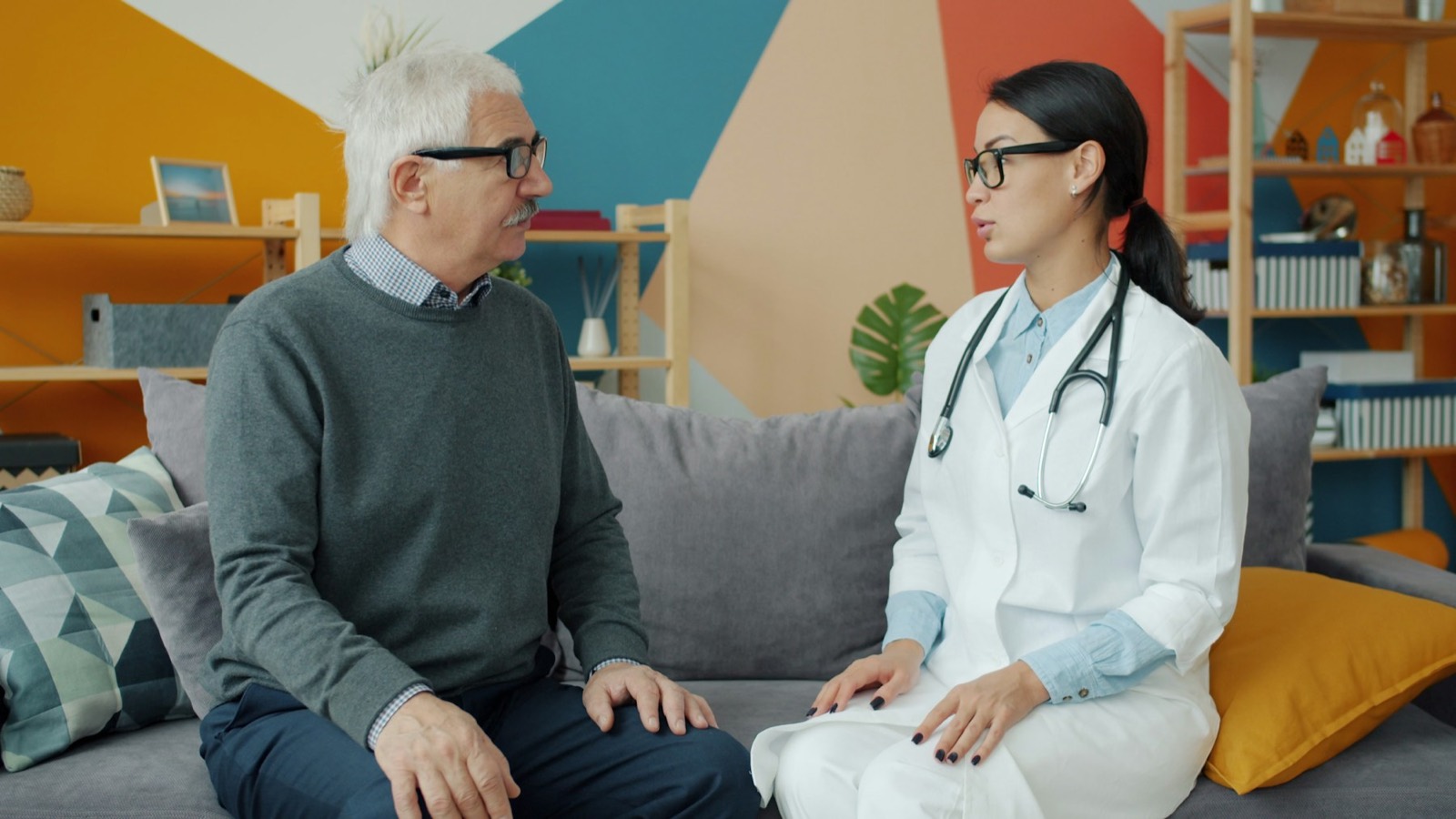 A doctor in a white coat talking with an older man seated on a sofa
