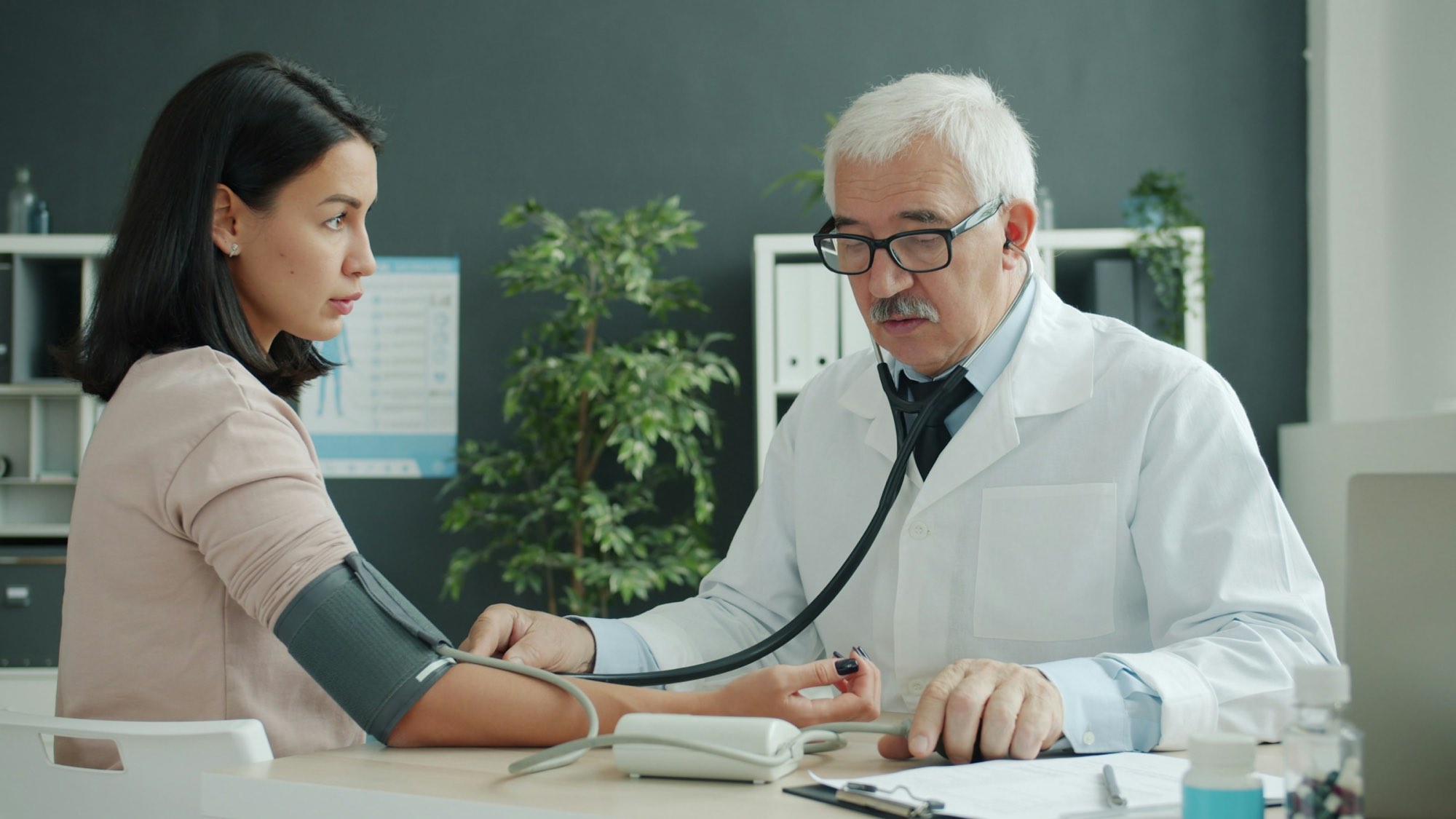 A doctor in a white coat checking a woman’s blood pressure at his desk