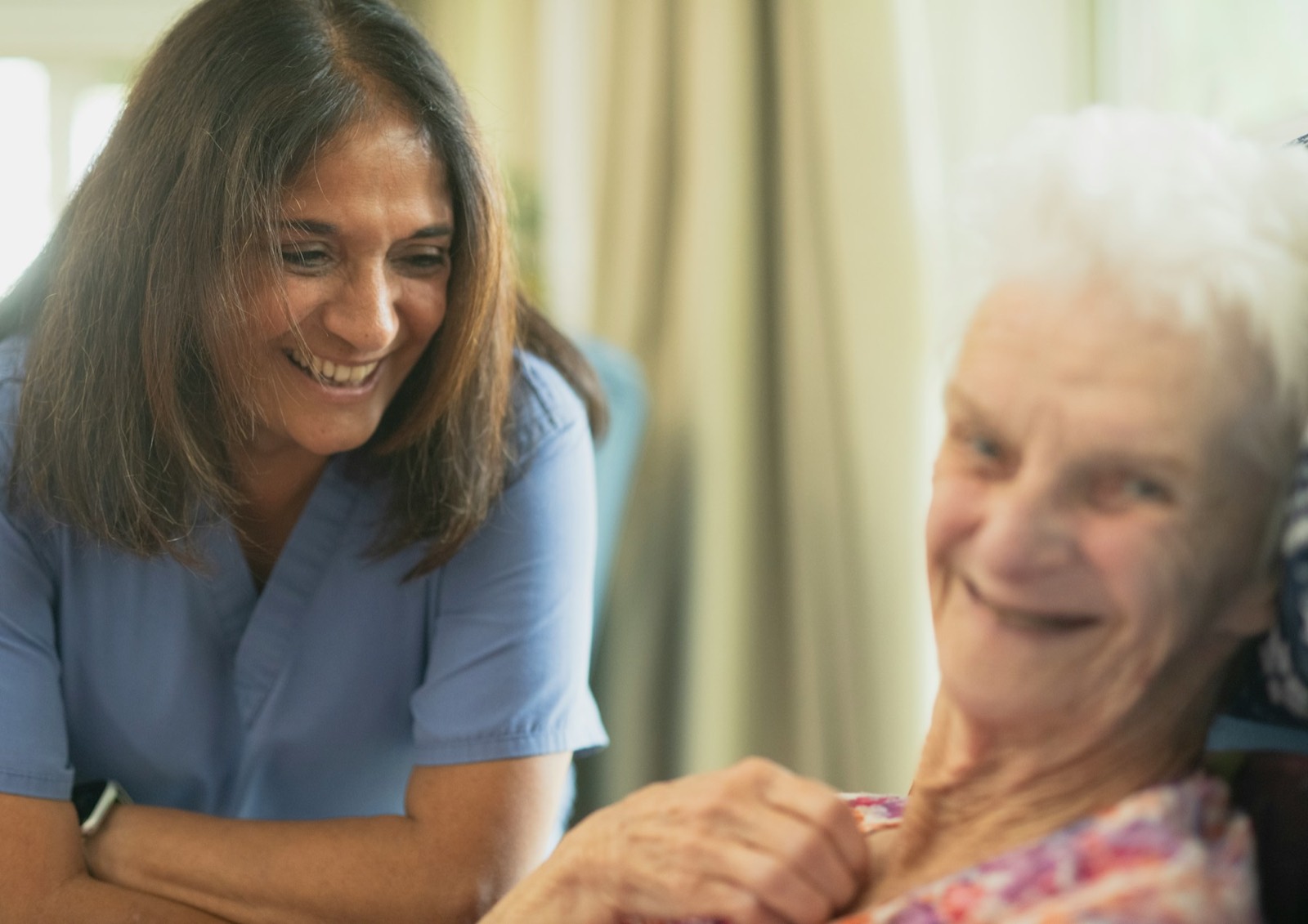 A nurse in blue scrubs chatting with an older woman in a care setting
